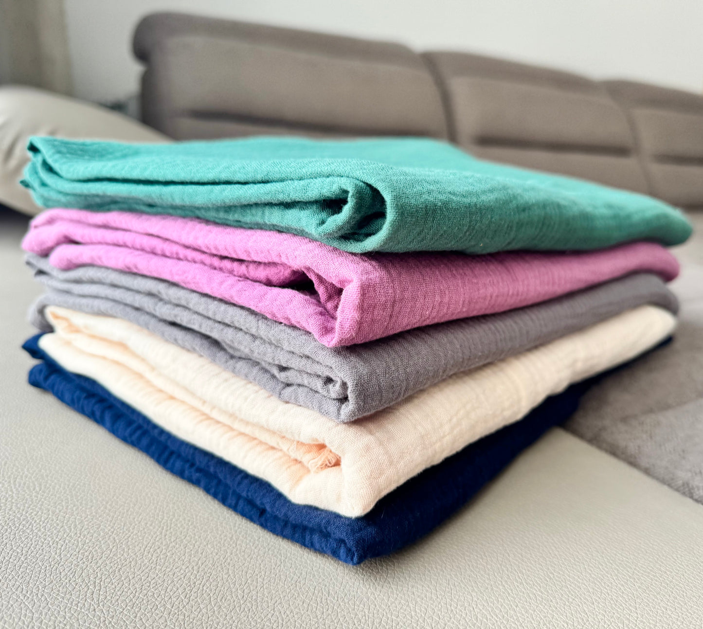 Stack of folded muslin cotton scarf wrap blankets in various colors on a sofa