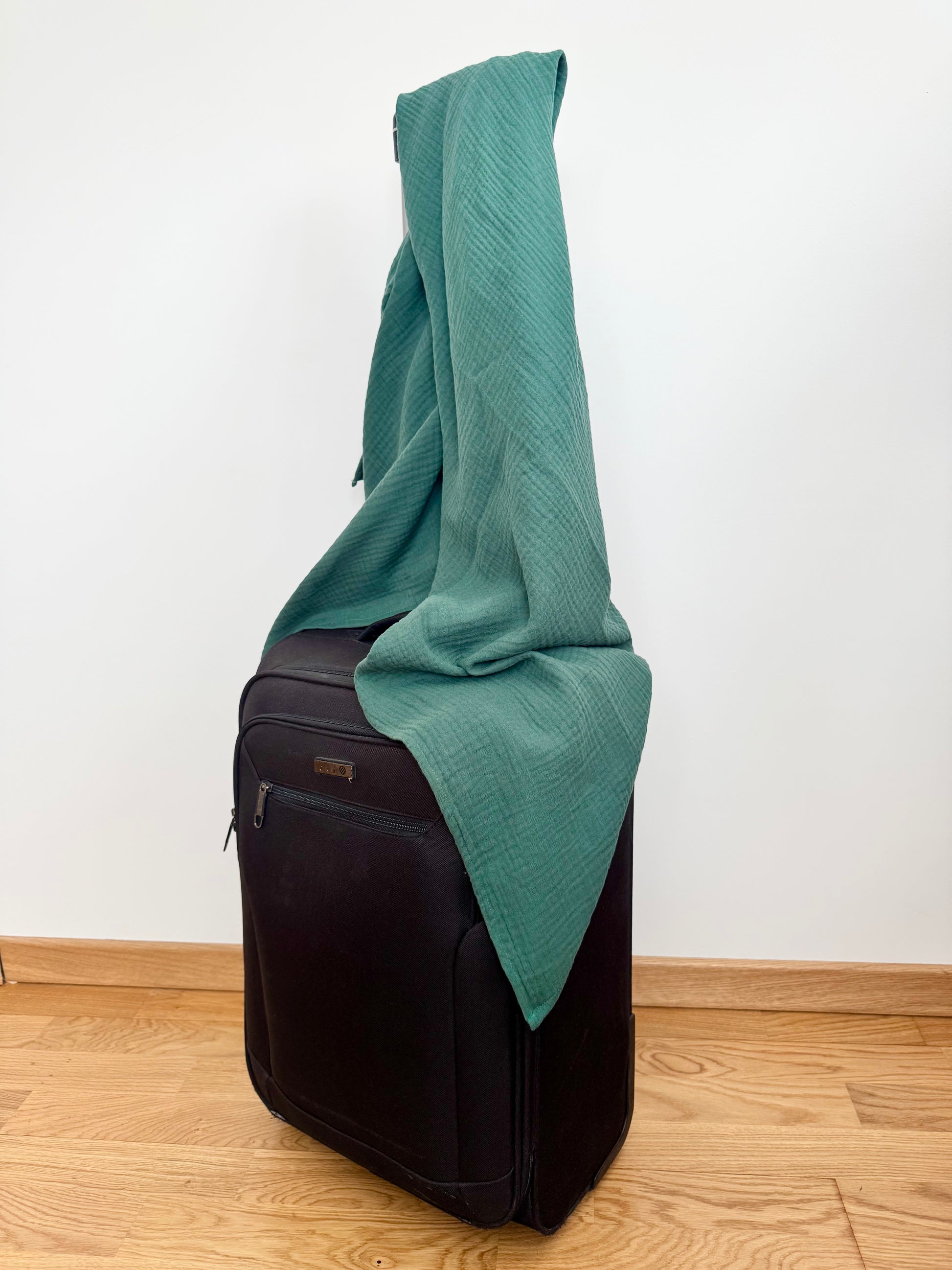 emerald Green scarf wrap blanket draped over a black suitcase on a wooden floor with a white wall background
